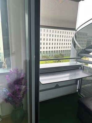 Ein Zimmer mit großem Fenster, grüner Teppich, kleinem Tisch und einer Zimmerpflanze.