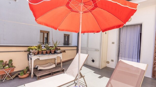 A balcony with a red umbrella, two lounge chairs, a small table with potted plants, and a door. - Vacation Home Schiavone Family (Matera)