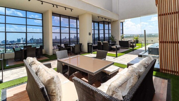 A rooftop lounge with wicker furniture, large windows offering city views, and a glass-top table. - Elite Residence View Palace Apartment (Nairobi)