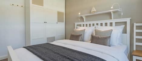 Quarto com cama, guarda-roupa branco e prateleira com abajur.