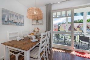 Un coin repas avec une table en bois, des chaises blanches, un luminaire suspendu en osier, un tableau représentant des voiliers et une vue sur un balcon avec des sièges d’extérieur.