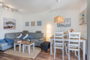 Un salon moderne avec un canapé gris, une table basse en bois, un ensemble de salle à manger blanc et des œuvres d’art accrochées aux murs.