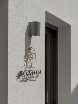 Un edificio con un logo y el nombre 'SIMEOS HOUSE' en la pared, una puerta y una lámpara fijada a la pared.
