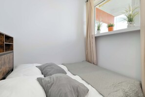 A small, neatly arranged bedroom with a bed, a wooden headboard, a window with curtains, and a shelf with potted plants. - Cozy apartment with a balcony | Pets welcome (Gdańsk)