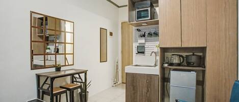 A compact room with a bed, a desk, a chair, a small kitchenette, and a mirror.