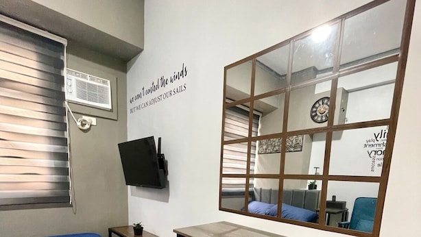 A compact room with a bed, a desk with two chairs, a TV mounted on the wall, and a large mirror reflecting the room. - Urban Deca Tower Mandaluyong-Cozy Stay (Mandaluyong City)
