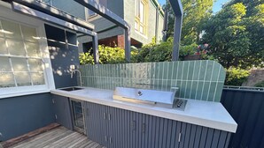 Et moderne udendørs køkken med indbygget vask og grill, træterrasse og en væg med grønne fliser.