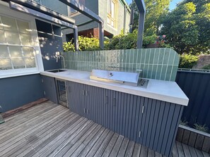 A modern outdoor kitchen with a built-in sink and grill, wooden deck flooring, and a green-tiled wall. - Boutique Garden Cottage | Pool & BBQ| Free Parking | Public Transport (Leichhardt)