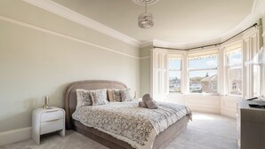 A bedroom with a large bed, a nightstand, a dresser, and a bay window with a view of buildings. - Spacious 4BR - Private driveway & Garden (Rutherglen)