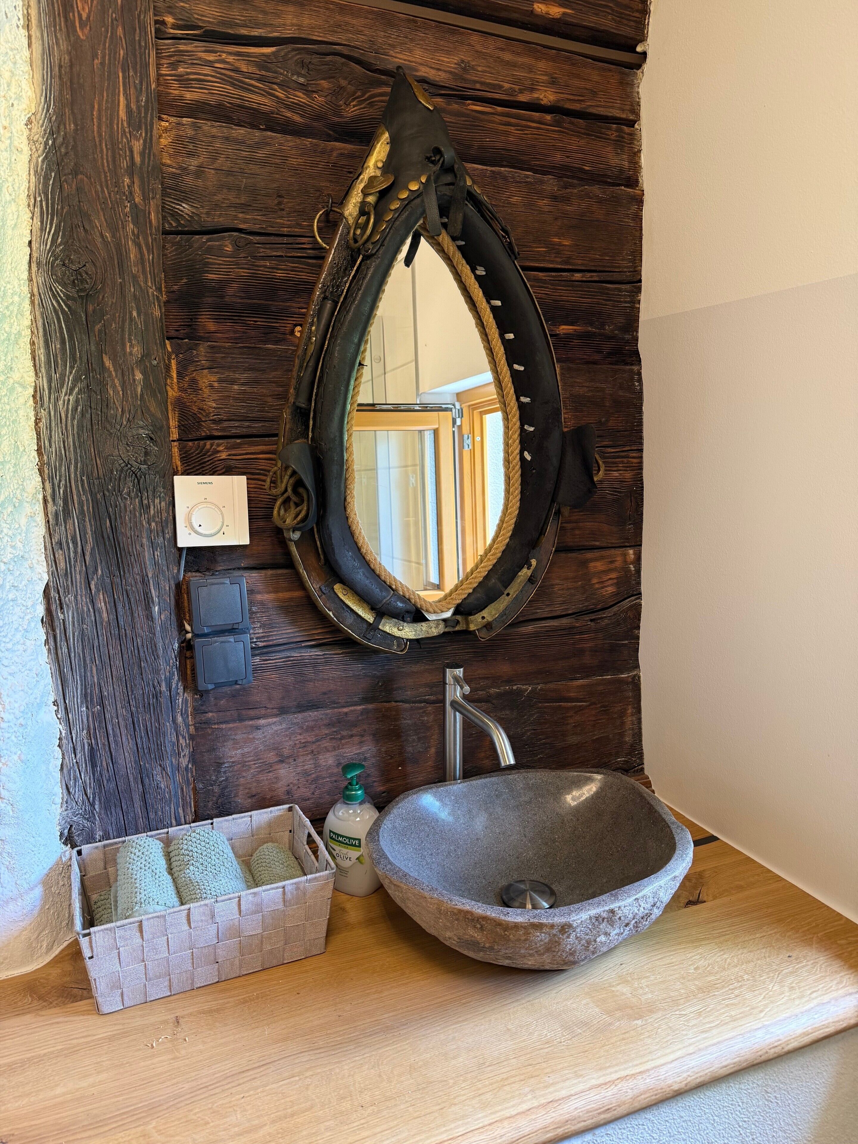 A rustic bathroom with a wooden wall, a stone sink, a mirror, a thermostat, and a soap dispenser.