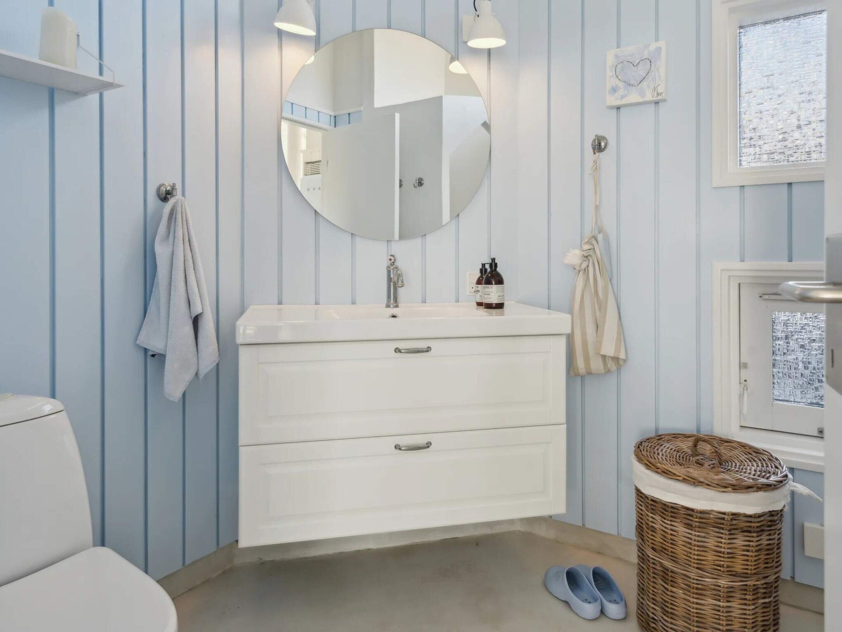 A bathroom with a white sink, a round mirror, a towel rack, a wicker basket, and a pair of slippers.