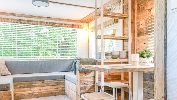 Interior acogedor de una cabaña con paredes de madera, un banco empotrado, una mesa con taburetes y una ventana con persianas.