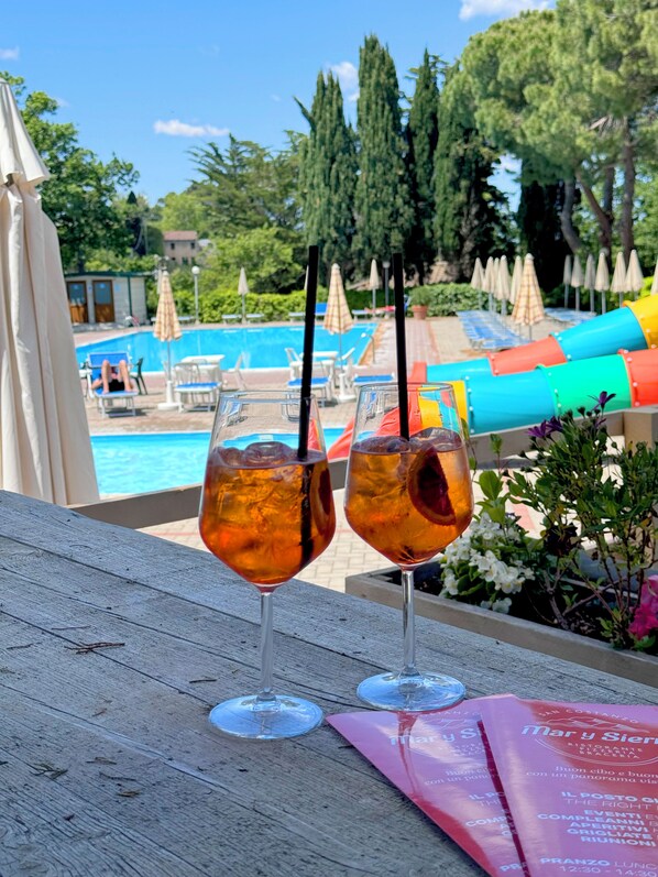 A poolside view with two glasses of amber beverage, a wooden table, and a menu on the right. - STOERbuiten Mar Y Sierra (Stacciola di San)
