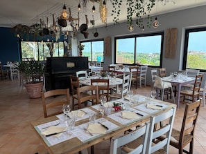 Un restaurant doté de nombreuses tables dressées pour le service, d’un piano et de grandes fenêtres offrant une vue sur la campagne.