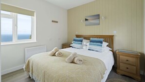 A bedroom with a bed, two bedside tables, a window with a view, a wall-mounted picture, and a radiator. - Kitticarn, Panoramic Sea Views, Garden, Parking (Sennen)