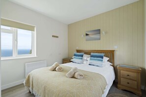 A bedroom with a bed, two bedside tables, a window with a view, a wall-mounted picture, and a radiator. - Kitticarn, Panoramic Sea Views, Garden, Parking (Sennen)