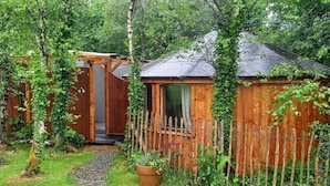 Una cabaña de madera con techo inclinado, rodeada por una valla de madera y abundante vegetación.