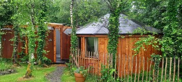 Una cabaña de madera con techo inclinado, rodeada por una valla de madera y abundante vegetación.
