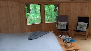 Una habitación de madera con una cama, dos sillas con almohadones con forma de mariposa, una mesita con un jarrón y dos ventanas con vistas al bosque.