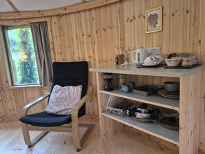 A wooden cabin interior with a rocking chair, a shelf with kitchenware, and a window with a view of greenery. - Unique Retreat, Sleeps 2 With Hot Tub & Pets Ok (Glengarriff)