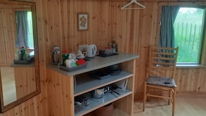 Una habitación de madera con un espejo, un estante con utensilios de cocina, una silla y una ventana con vistas a la vegetación.