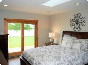 A bedroom with a bed, a nightstand, a lamp, a decorative wall piece, and a sliding glass door leading to a green outdoor area. - Lake St. Helen Retreat with Pontoon, Kayaks, Hot Tub & Easy ORV Trail Access (St. Helen)