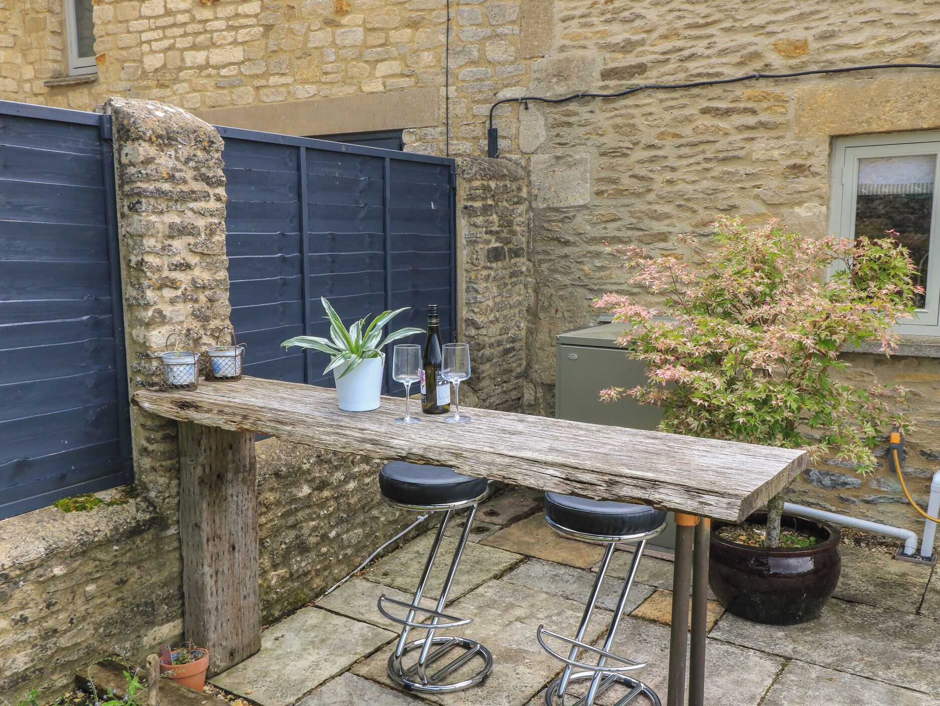 Un espace bar extérieur de style rustique, avec un mur en pierre en arrière-plan, un comptoir en bois, deux tabourets, une plante en pot et une bouteille de vin.