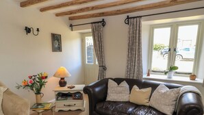 A living room with a leather sofa, a small table with a lamp, a vase of flowers, and a window with curtains. - 2 The Gardens (Lechlade)