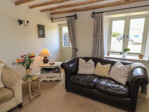 A living room with a leather sofa, a small table with a lamp, a vase of flowers, and a window with curtains. - 2 The Gardens (Lechlade)