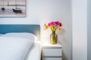 Ein ordentlich bezogenes Bett mit blauem Kopfteil, ein weißer Nachttisch mit Blumenvase und ein Bild von Booten an der Wand.