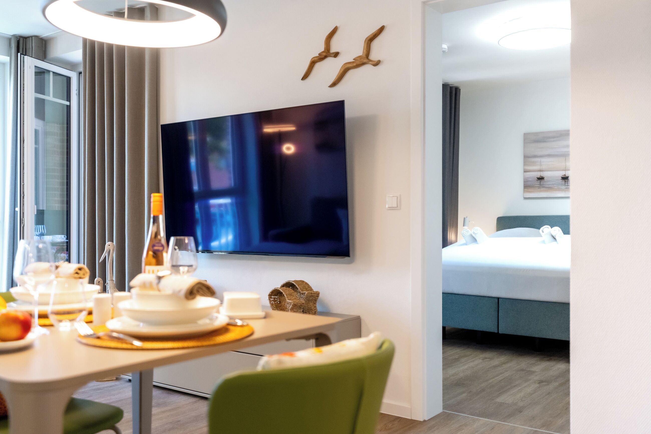 A modern hotel room with a dining area, a flat-screen TV, a bed with a blue headboard, and a painting on the wall.