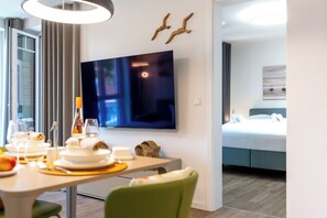 Quarto de hotel moderno com área de jantar, televisão de tela plana, cama com cabeceira azul e um quadro na parede.