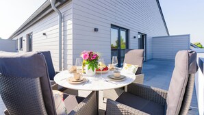 Een modern huis met een schuin dak, een balkon met buitenmeubilair en een eettafel gedekt voor twee personen.
