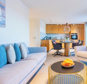 Ein modernes Wohnzimmer mit einer Couch, einem runden Couchtisch mit Obst und einer Küche im Hintergrund.