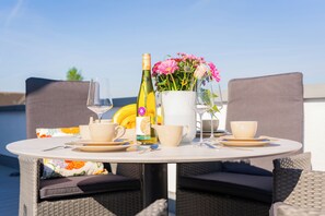 Ein runder Tisch mit weißer Tischdecke, zwei Korbstühle, einer Weinflasche, einem Blumenkrug und einem Glas Wasser.
