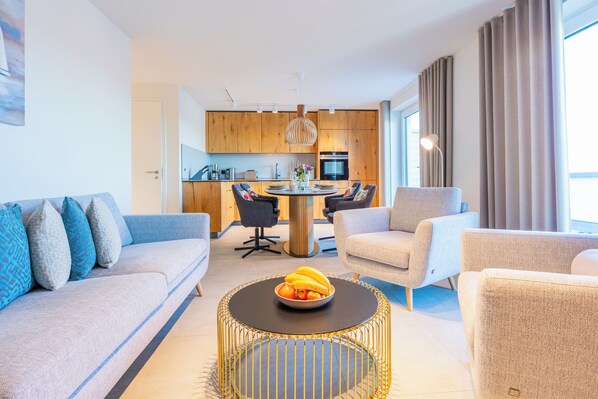 Ein modernes Wohnzimmer mit einer Couch, Sesseln, einem Couchtisch mit Obst und einer Küche im Hintergrund.