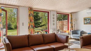 A room with a brown leather sofa, a round wooden table with a banana and a plate, and large windows with curtains. - 5 Star Holiday Home in Gedser (Gedser)