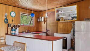 A kitchen with wooden cabinets, a built-in oven, a refrigerator, a window with a view, and a dining area with a table and chairs. - 5 Star Holiday Home in Gedser (Gedser)