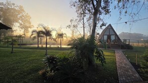 A house with a brick pathway leading to a wooden deck, surrounded by lush greenery and trees. - A retreat at Chalé A-Frame Parobé, with a lake, fire pit, and grill. (Poço Fundo)