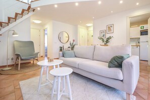 Ein modernes Wohnzimmer mit einer Couch, zwei weißen Hockern, einem Sessel, einem runden Spiegel und einer Treppe.