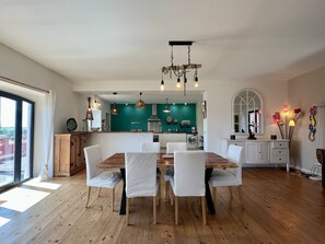 A spacious dining area with a wooden table and white chairs, a kitchen with teal walls, and a large window providing a view.