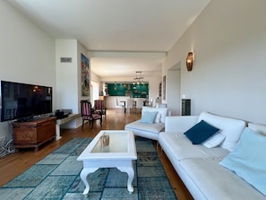 A living room with a television, sofa, coffee table, and chairs. There is a dining area in the background with a green wall and a kitchen area with a white countertop. - Villa Tagus Gardians (Trafaria)