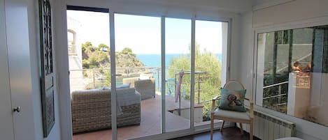 Una habitación con un gran ventanal corredera que da a un balcón con mobiliario exterior y vistas al mar.
