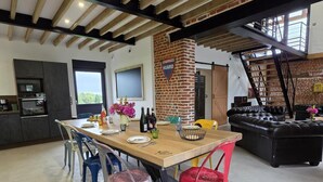 A modern kitchen with built-in appliances, a dining area with colorful chairs, and a brick fireplace. - Cottage with air conditioning, garden - 6 guests (Taisnières-en-Thiérache)