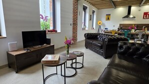 A modern living room with a TV, leather sofa, and a small table with a Monopoly game box. - Cottage with air conditioning, garden - 6 guests (Taisnières-en-Thiérache)