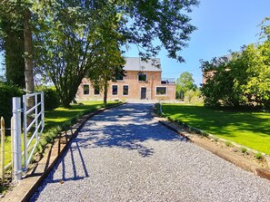 A gravel pathway leading to a brick building with multiple windows and a door, surrounded by greenery and trees.