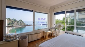 Une chambre avec une grande fenêtre donnant sur la côte, un lit, un bureau et un ventilateur sur pied.