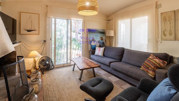 Un salon comprenant un canapé, une table basse en bois, un luminaire suspendu en osier, un ventilateur et une grande fenêtre donnant sur des arbres.