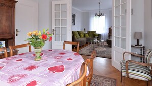 Un coin repas avec une table dressée pour deux personnes, un vase de fleurs et une vue sur un salon agrémenté d’un canapé et d’un lustre.
