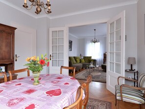 Un coin repas avec une table dressĂ©e pour deux personnes, un vase de fleurs et une vue sur un salon agrĂ©mentĂ© dâun canapĂ© et dâun lustre.